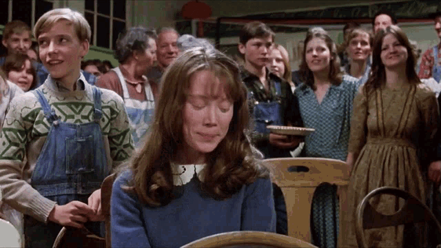 a group of people are gathered in a room including a girl in overalls