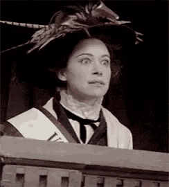 a woman wearing a hat and a sash is standing on a balcony and making a funny face .