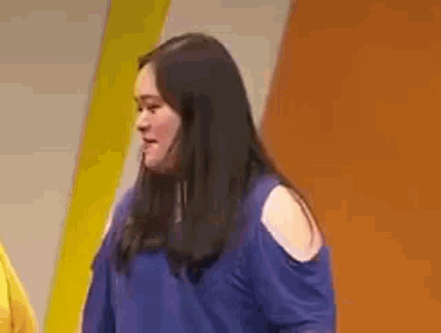 a woman in a blue off the shoulder top is standing in front of a yellow wall .