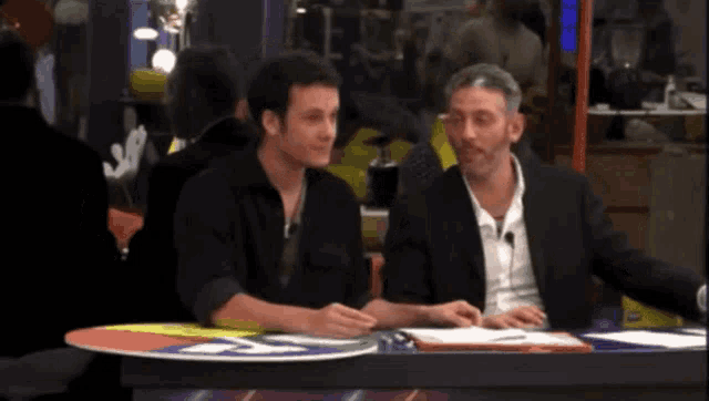 two men are sitting at a table with a sign that says ' sd '