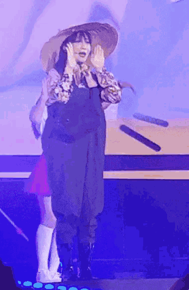 a woman wearing a hat and overalls is standing on a stage in front of a blue light .