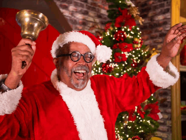 a man in a santa suit is holding a bell in front of a christmas tree