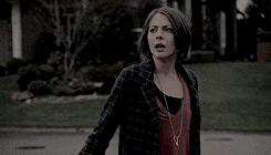 a woman in a plaid jacket is standing in front of a house with a surprised look on her face .