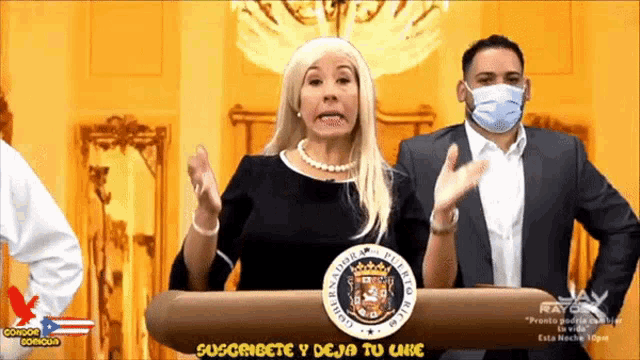a woman stands behind a podium that says suscribete y deja tu like