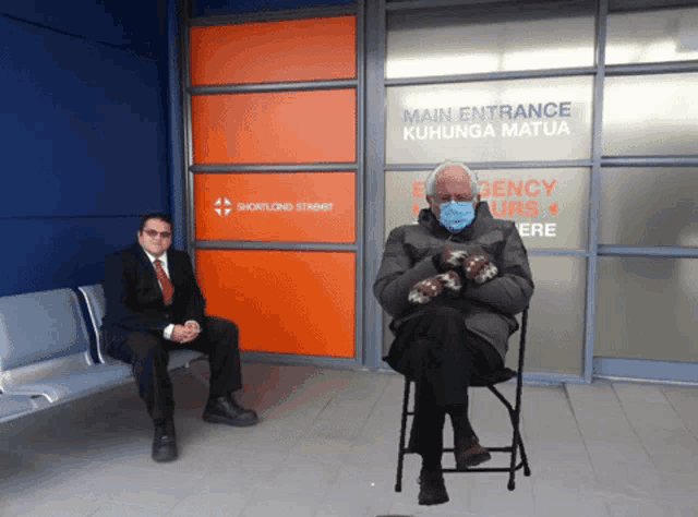 a man in a suit sits next to a man in a chair in front of a sign that says main entrance