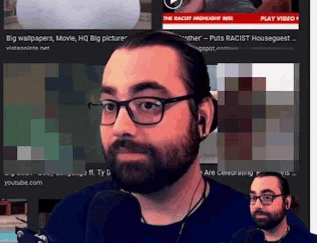 a man with glasses and a beard is talking into a microphone in front of a play video screen
