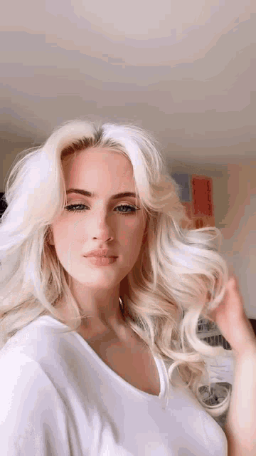 a woman with blonde hair and blue eyes is wearing a white shirt and holding her hair .