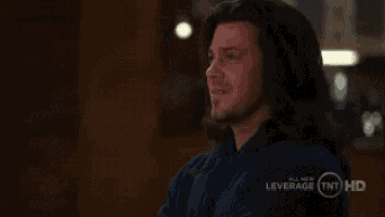 a man with long hair and a beard is sitting in a dark room and looking at the camera .