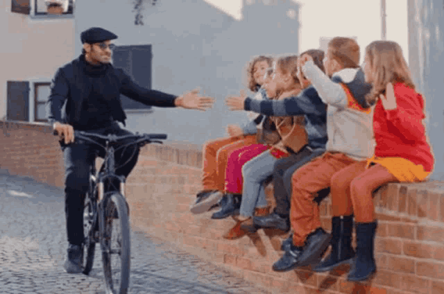 a man on a bike is talking to a group of children who are sitting on a brick wall