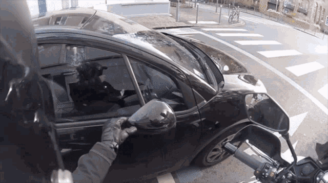 a person holding a motorcycle 's rear view mirror while a car is parked on the side of the road