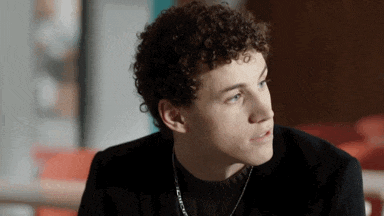 a young man with curly hair and blue eyes is wearing a black jacket and a necklace .