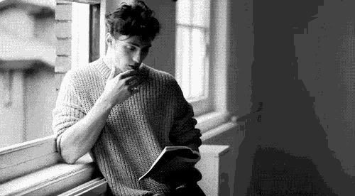 a man is sitting on a window sill reading a book while drinking coffee .
