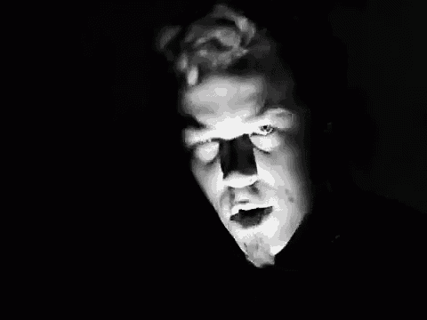 a black and white photo of a man with a beard looking at the camera in the dark .