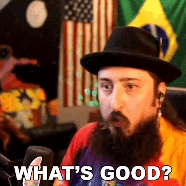 a man with a beard wearing a hat says " what 's good " in front of a microphone