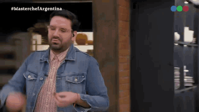 a man wearing a denim jacket and glasses is dancing in front of a brick wall and a shelf with plates on it