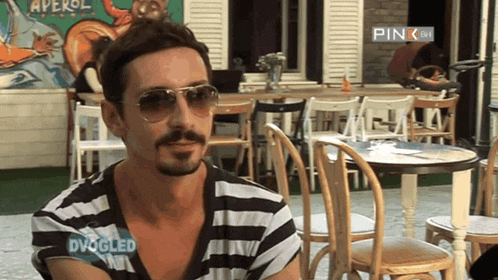 a man wearing sunglasses and a shirt that says dvogled sits in front of tables and chairs