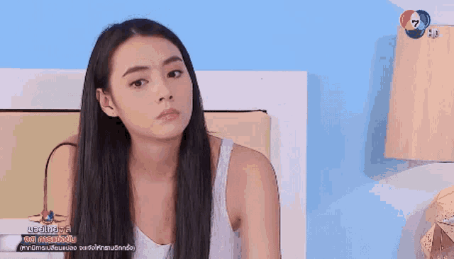 a woman in a white tank top is sitting on a bed in front of a blue wall and a lamp