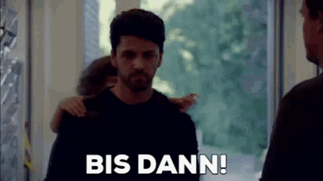 a man with his arm around another man 's shoulder is standing in front of a window and says bis dann .