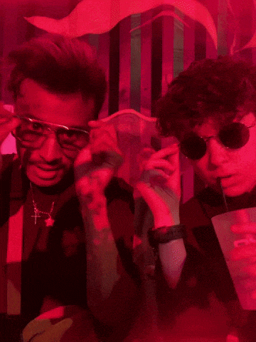 two men wearing sunglasses and suspenders pose for a picture in a dark room