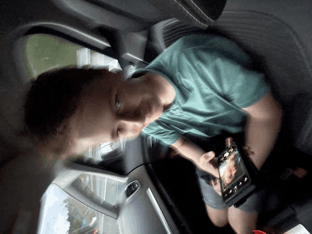 a boy in a green shirt sits in the back seat of a car holding a tablet