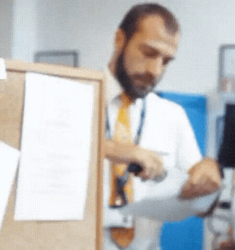a man with a beard is standing in front of a bulletin board looking at a piece of paper .