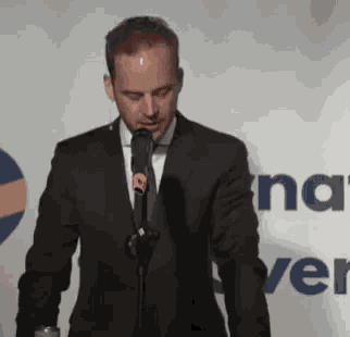 a man in a suit is speaking into a microphone in front of a wall that says na