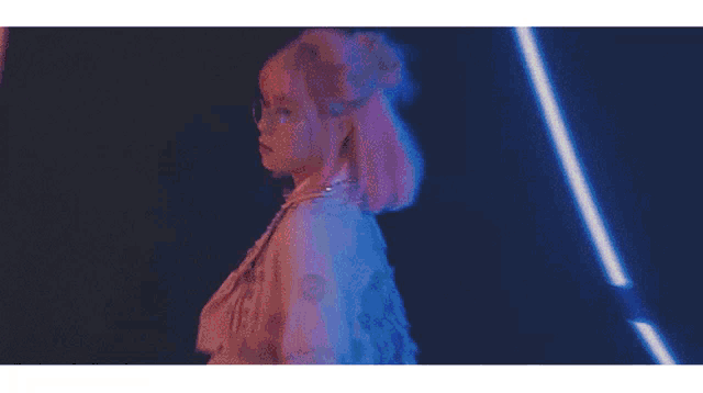 a woman with pink hair and glasses stands in front of a neon light