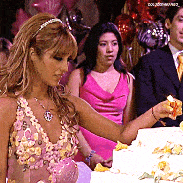 a woman in a pink dress is cutting a cake with a peace sign on her necklace