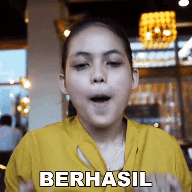 a girl in a yellow shirt is making a funny face and the word berhasil is on her shirt