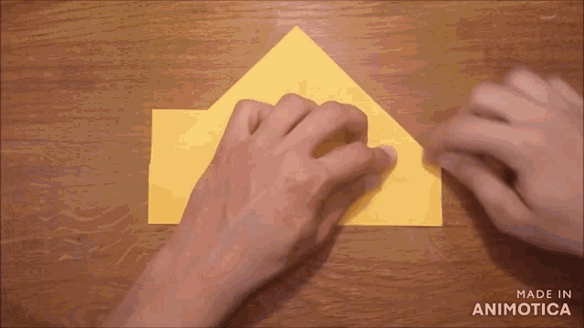 a person folding a piece of yellow paper on a wooden table with the words made in animotica below them