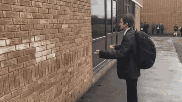 a young man with a backpack is standing next to a brick wall .