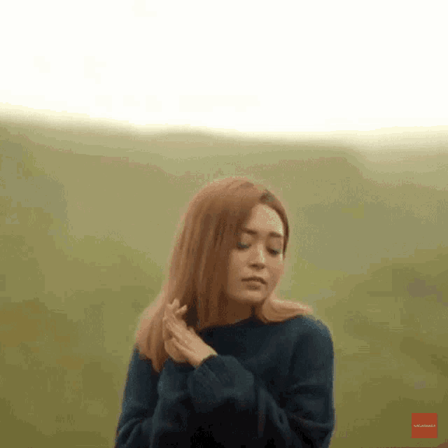 a woman in a blue sweater is standing in a field with her head down