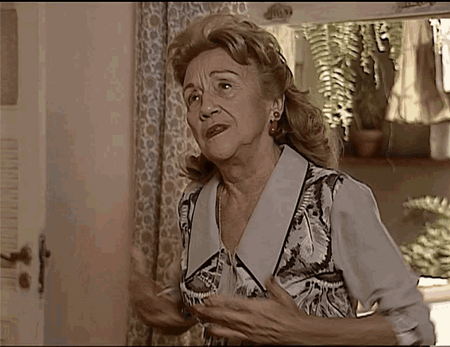 an elderly woman in a floral shirt stands in front of a window