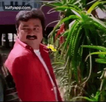 a man in a red shirt with a mustache is standing in front of a plant .
