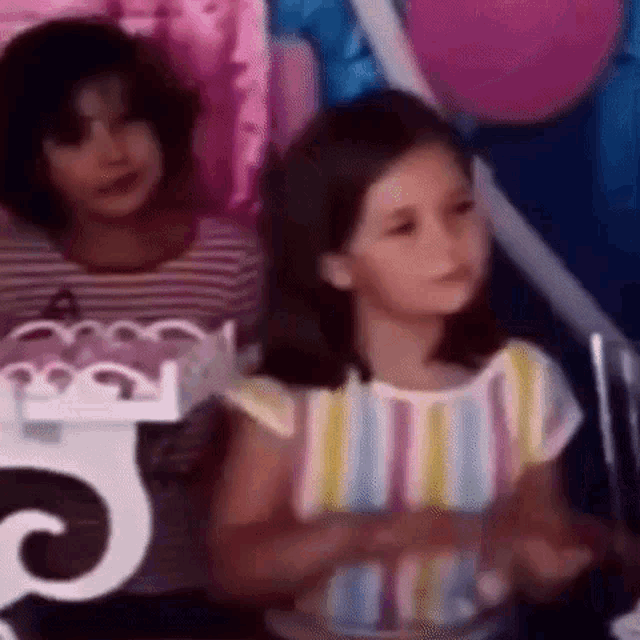 two little girls are sitting next to each other at a birthday party . one of the girls is wearing a striped shirt .