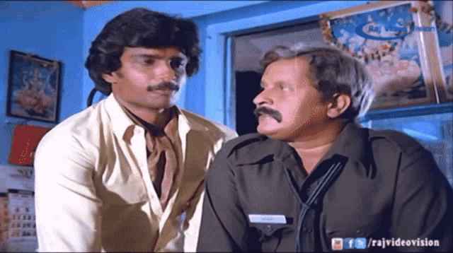 two men are standing next to each other in front of a blue wall and a sign that says raj videovision