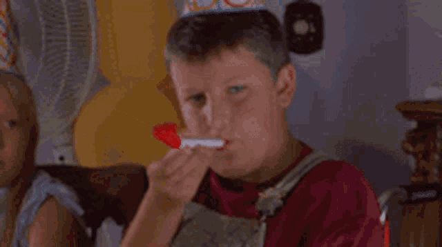 a boy wearing a party hat is blowing a party horn while a girl looks on .