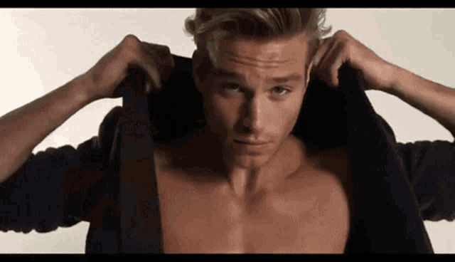 a shirtless man is taking off his jacket and adjusting his collar .