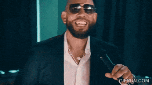 a man wearing sunglasses and a suit is holding a microphone and smiling .