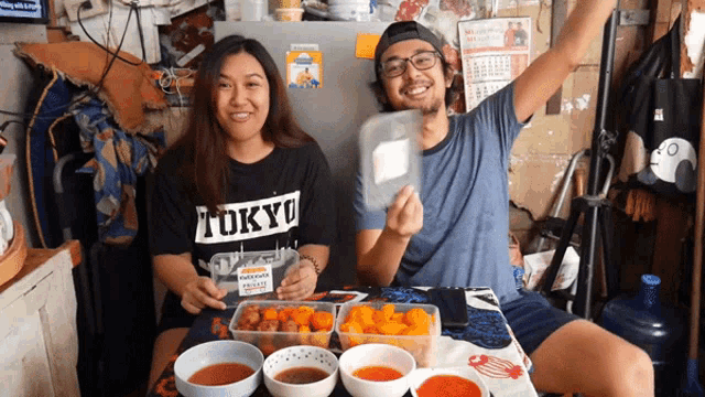 a man and a woman are sitting at a table with bowls of food and the woman is wearing a tokyo t-shirt