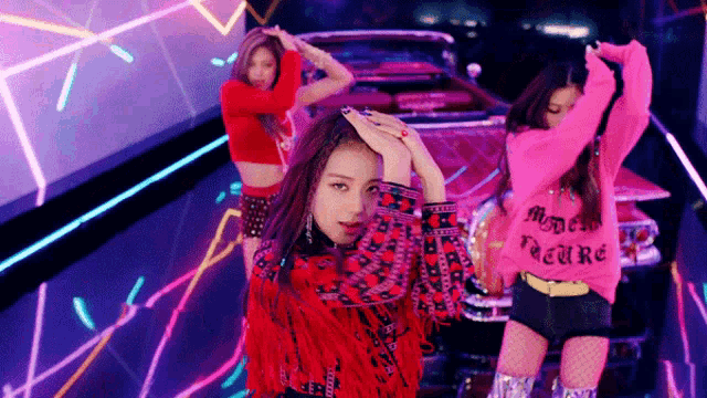 three women are dancing in front of a car and one of them is wearing a pink sweater that says mode nature