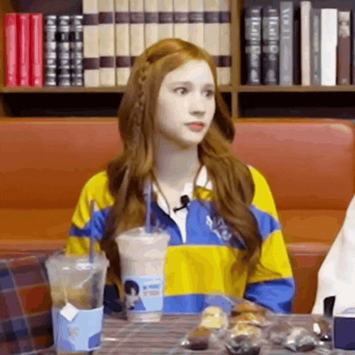 a young girl is sitting at a table with a cup of coffee and a bookshelf in the background .