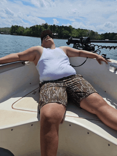 a man in a white tank top and camo shorts is laying on a boat with his arms outstretched