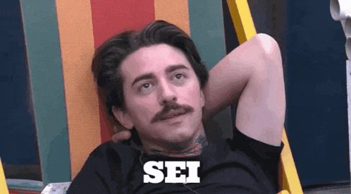 a man with a mustache is sitting in a chair and the word sei is on his shirt