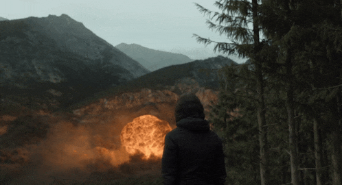 a person in a hooded jacket stands in front of a mountain with a large explosion in the distance