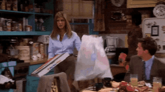 a woman in a blue shirt is holding a pizza box while a man in a suit sits at a table with a bag