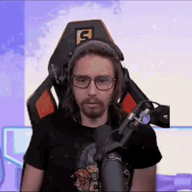 a man wearing glasses and headphones is sitting in front of a microphone in a gaming chair .