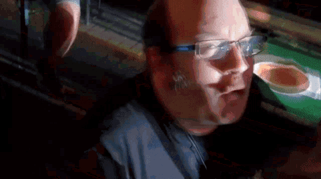 a man wearing glasses is sitting in a dark room with a bowl of food in the background .