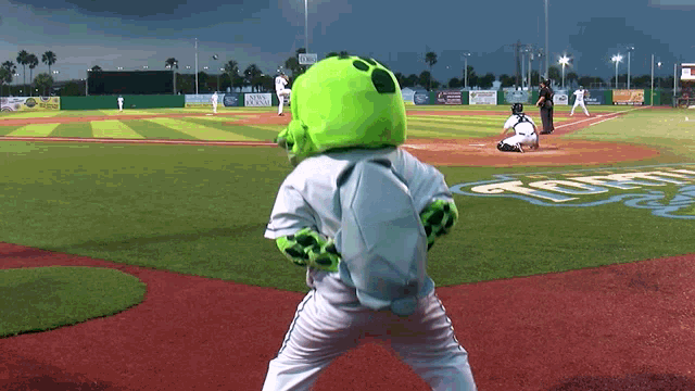 a green alien mascot stands on a baseball field watching a game