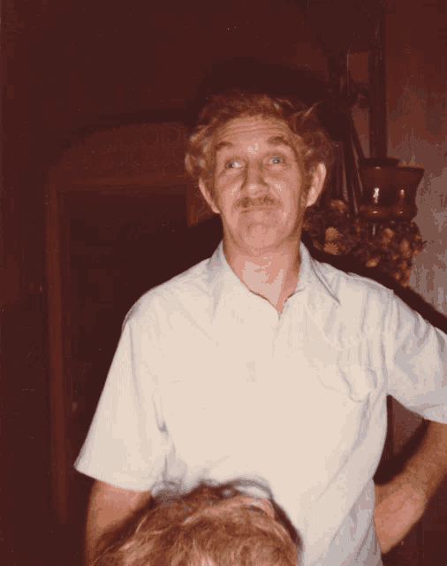 a man with a mustache wearing a white shirt stands in a dark room
